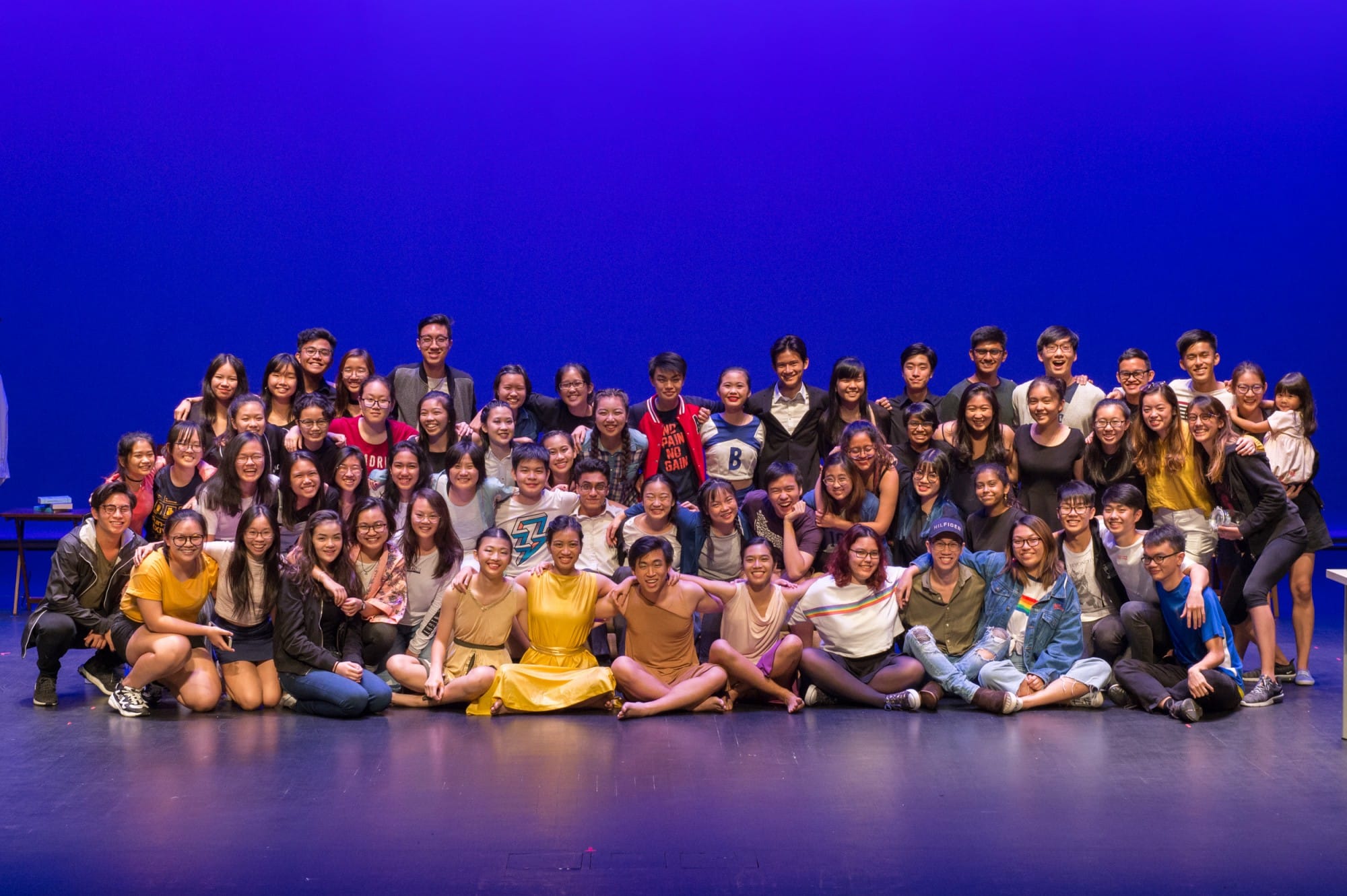 A large group of young people posing together on a stage with a bright blue backdrop, smiling at the camera.